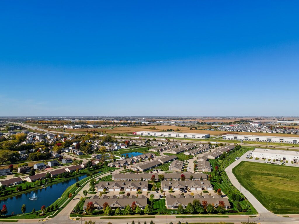 Aerial view of a suburban neighborhood with houses, ponds, roads, green spaces, and industrial buildings visible in the distance under a clear blue sky.
