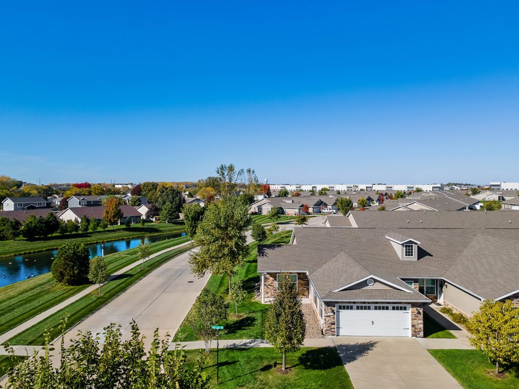 A suburban neighborhood with single-story houses, a pond, green lawns, and tree-lined streets under a clear blue sky.