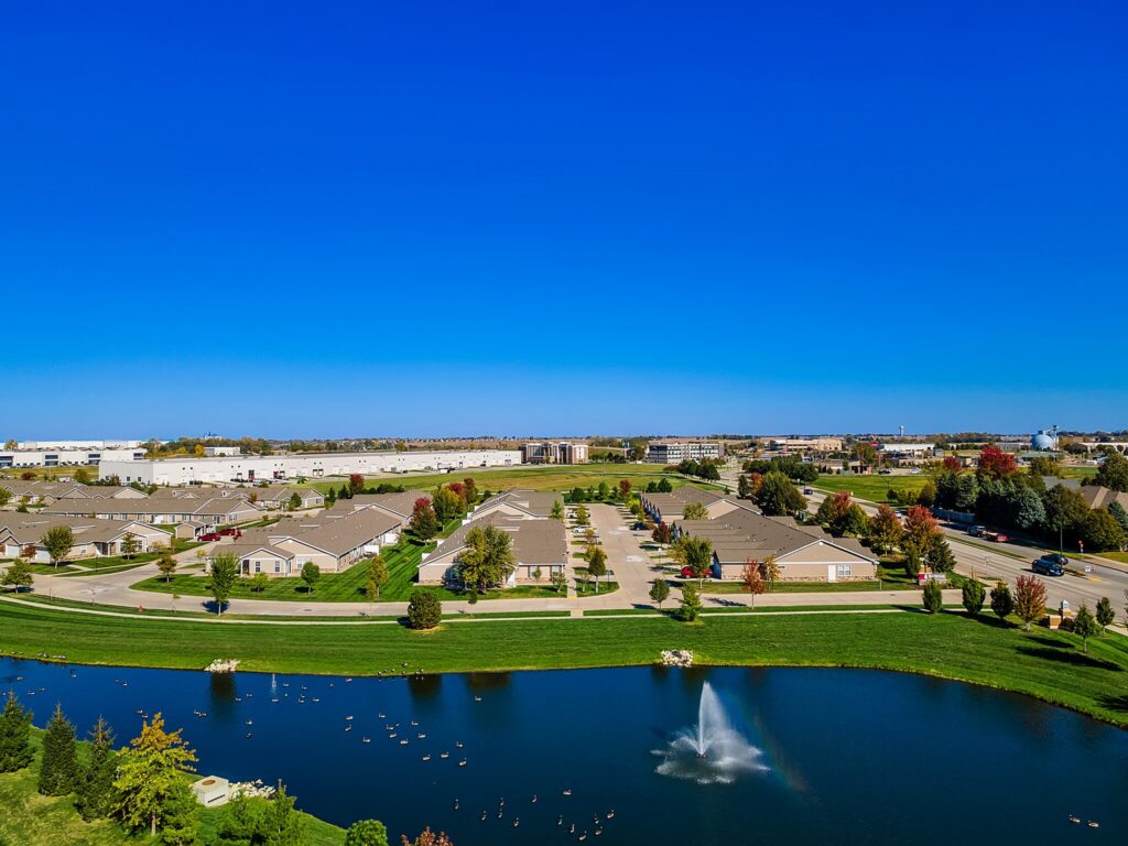 Aerial view of a residential neighborhood with a small lake, fountain, and several single-story homes under a clear blue sky.