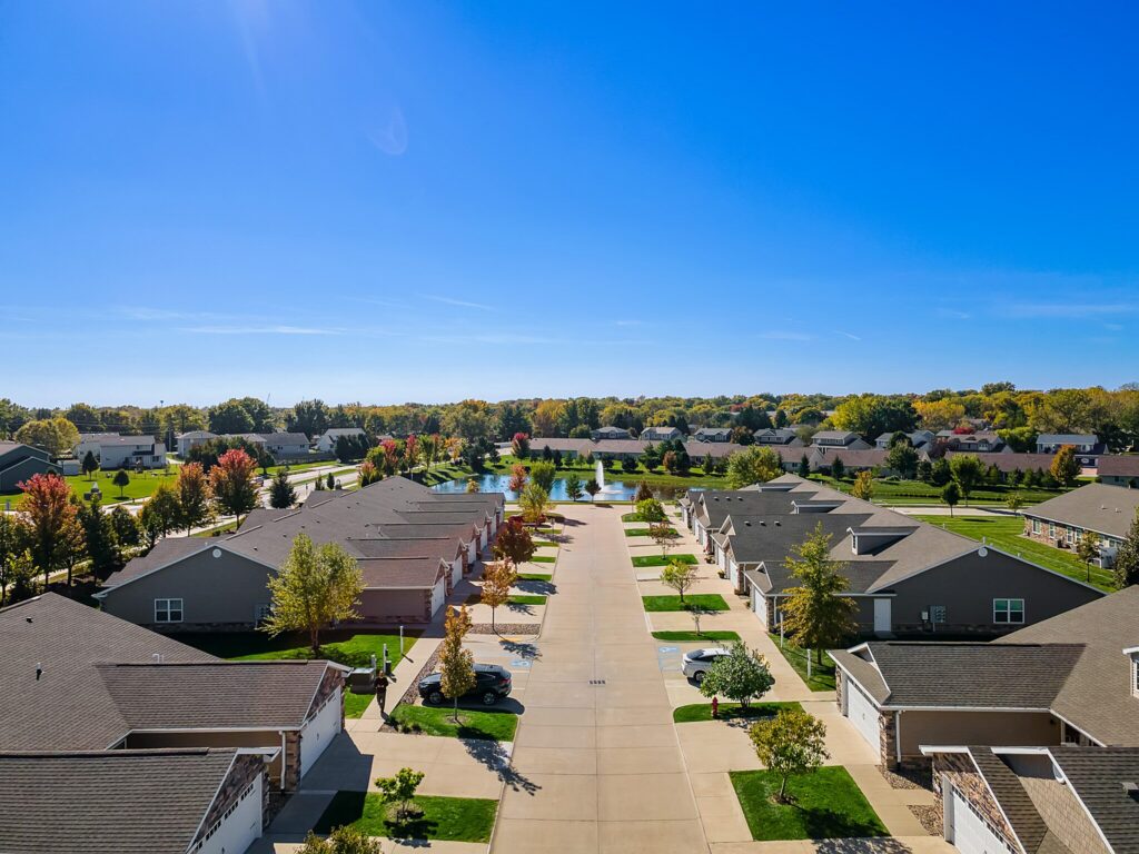 Aerial view of a suburban neighborhood with single-story houses, driveways, cars parked, green lawns, and trees under a clear blue sky.