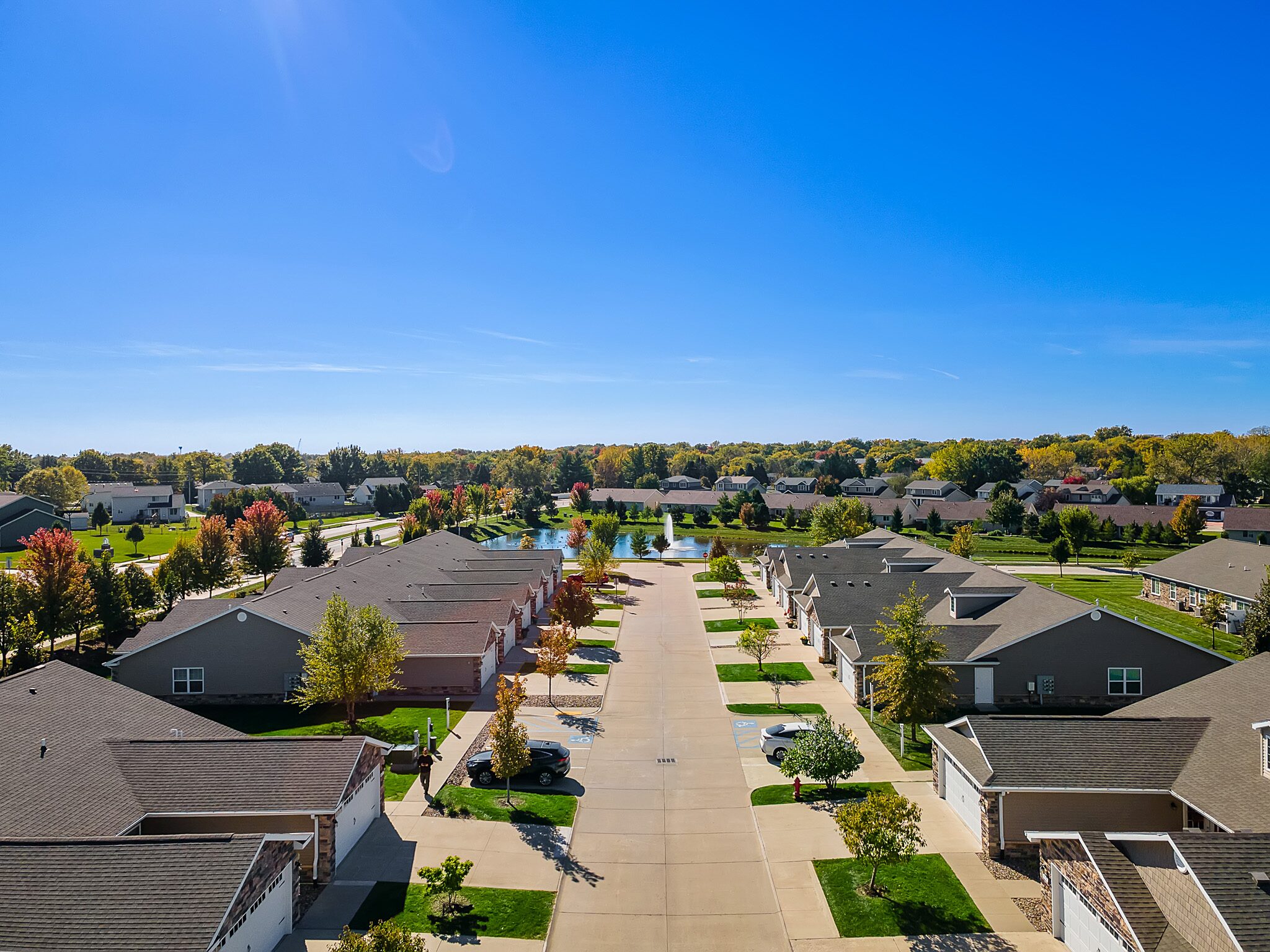 Aerial view of a suburban neighborhood with single-story houses, driveways, cars parked, green lawns, and trees under a clear blue sky.