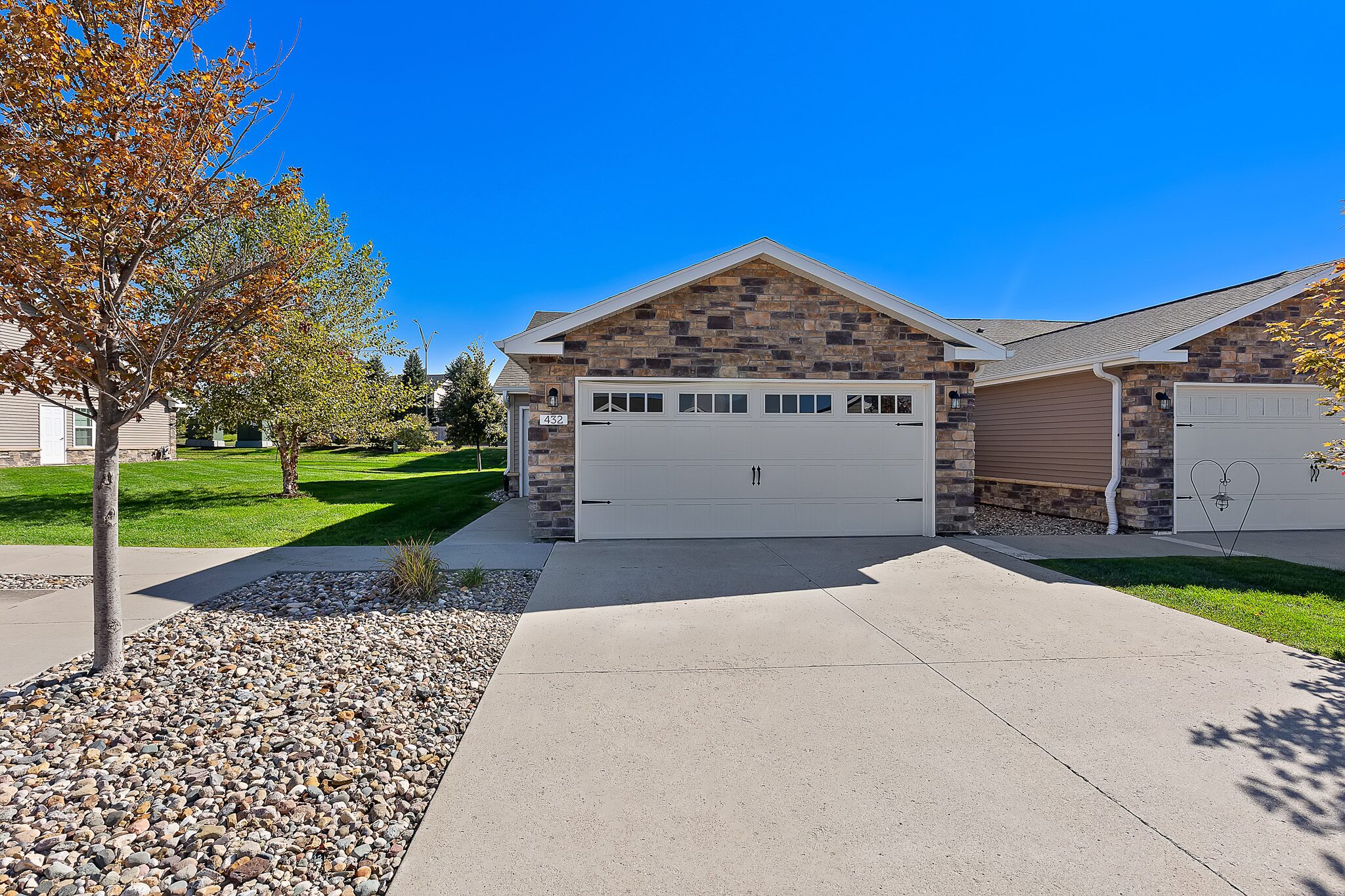 Single-story house with a stone facade, double garage, concrete driveway, and landscaped front yard with rocks, trees, and green grass under a clear blue sky.