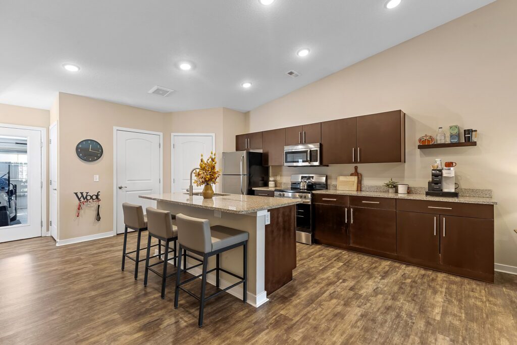 Modern kitchen with brown cabinets, stainless steel appliances, granite island with three barstools, and wooden flooring. Decorative items and a wall clock are visible.