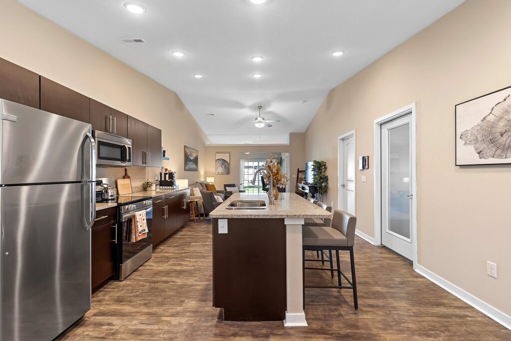 Modern kitchen with dark cabinets, stainless steel appliances, center island with seating, and wood flooring, leading to a living area in a neutral-toned open-plan apartment.