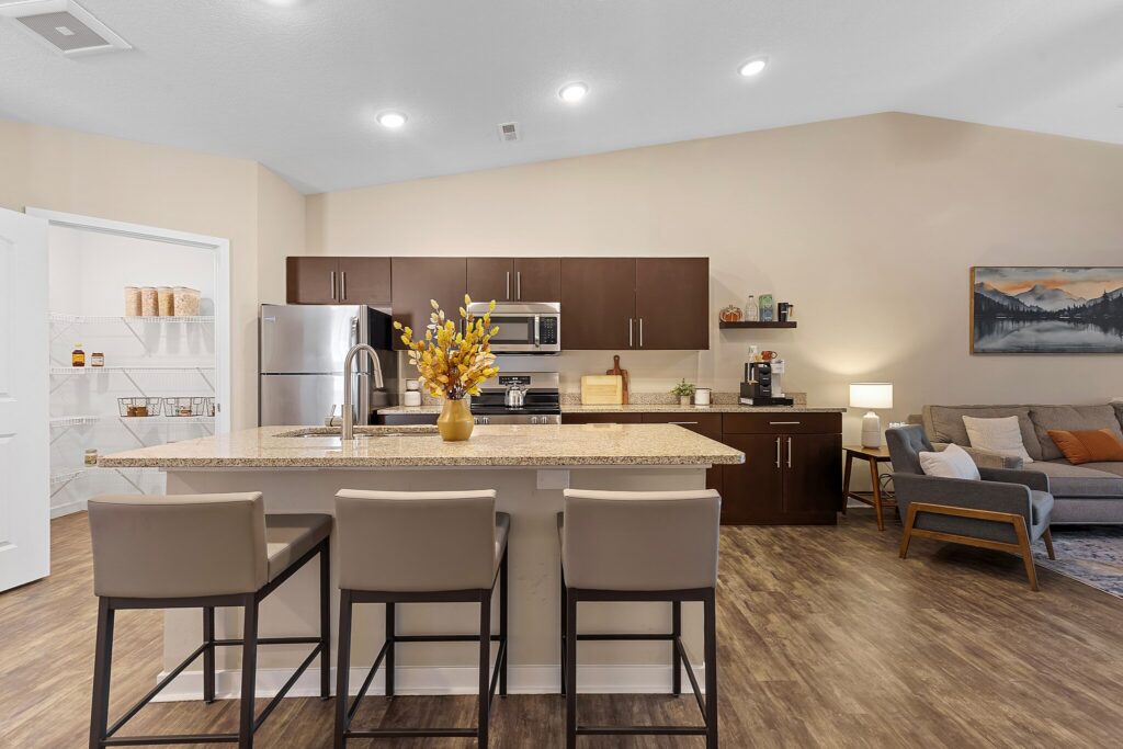 Modern kitchen with brown cabinets, stainless steel appliances, a granite island with three chairs, and an open pantry; living room area with sofa and artwork visible in the background.
