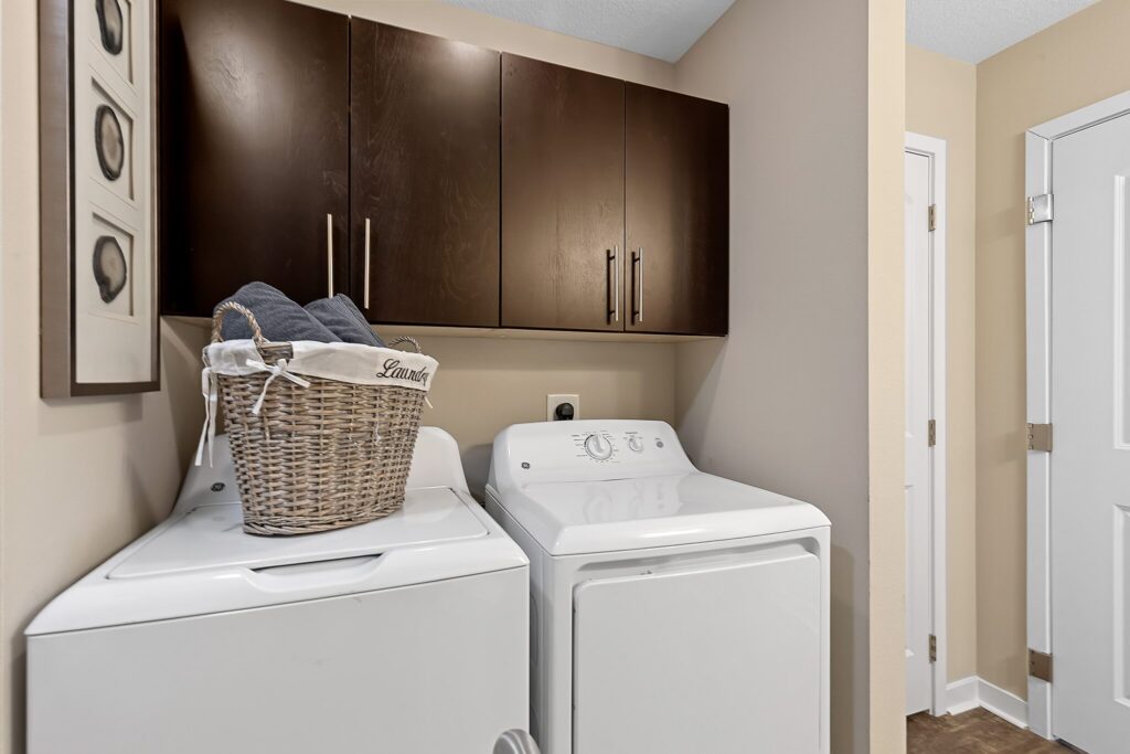 Laundry room with a washer and dryer, a wicker laundry basket on top, dark cabinets above, and a closed door to the right.