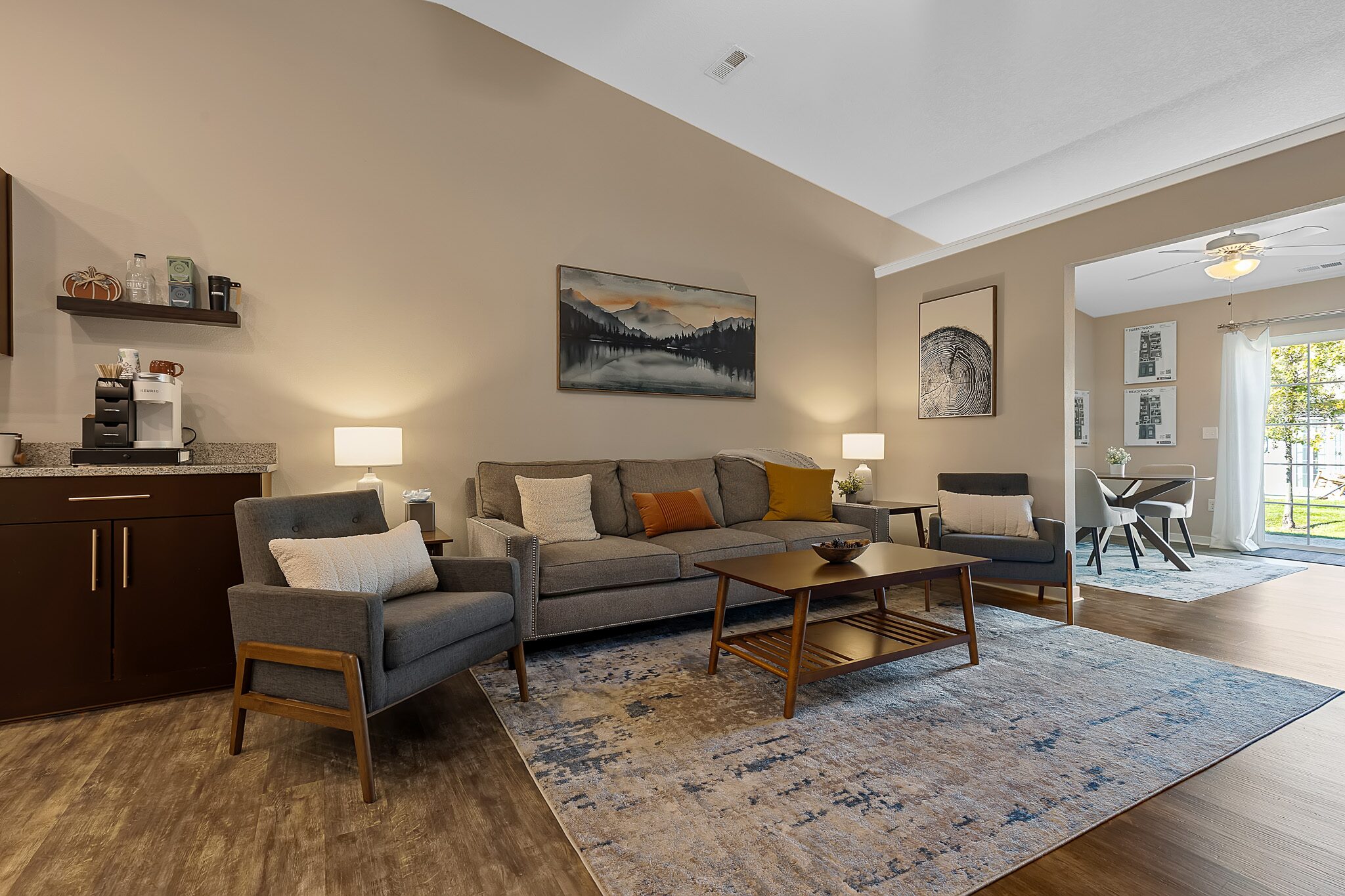 Modern living room with a gray sofa, two armchairs, a wooden coffee table, and neutral decor; adjacent dining area visible with large windows and natural light.