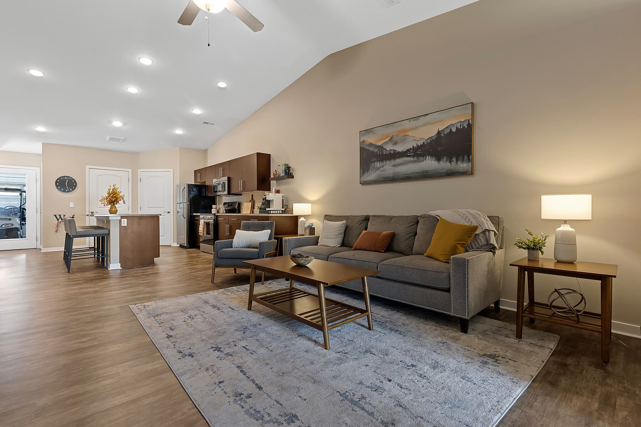 Open-concept living room and kitchen with gray sofa, wooden coffee table, dining island, and warm lighting; landscape artwork and neutral decor throughout.