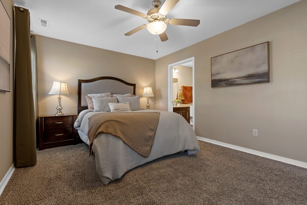 Bedroom with a neatly made bed, two bedside tables with lamps, ceiling fan, carpeted floor, and open doorway to a bathroom.