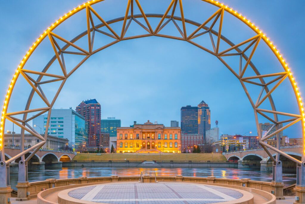 A city skyline with a river, modern buildings, and a historic building framed by a lit metal arch structure at dusk.