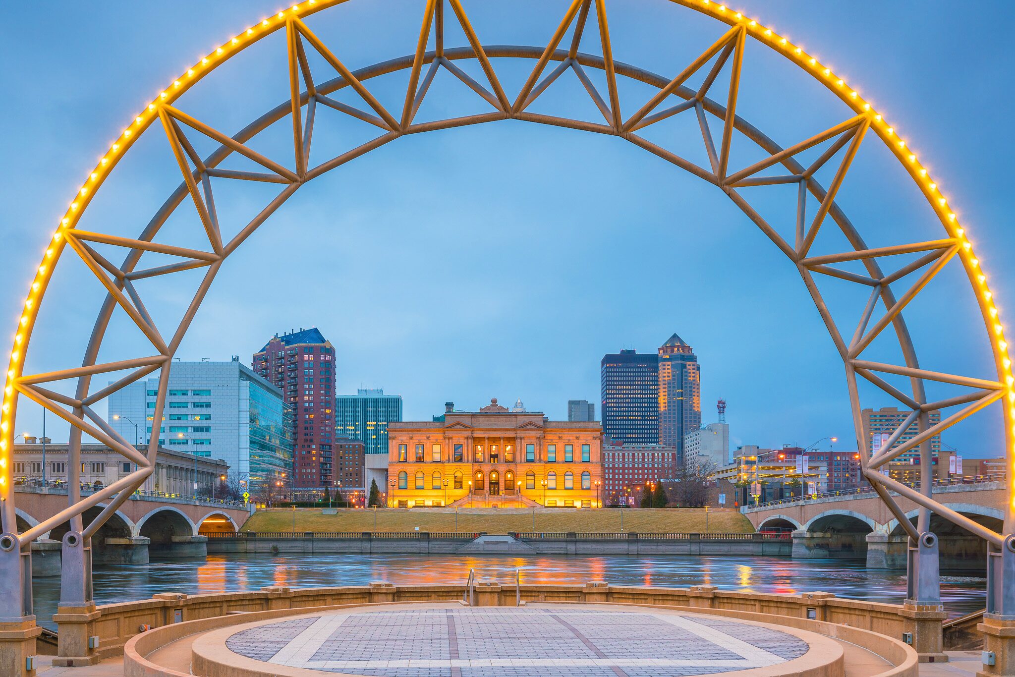 A city skyline with a river, modern buildings, and a historic building framed by a lit metal arch structure at dusk.