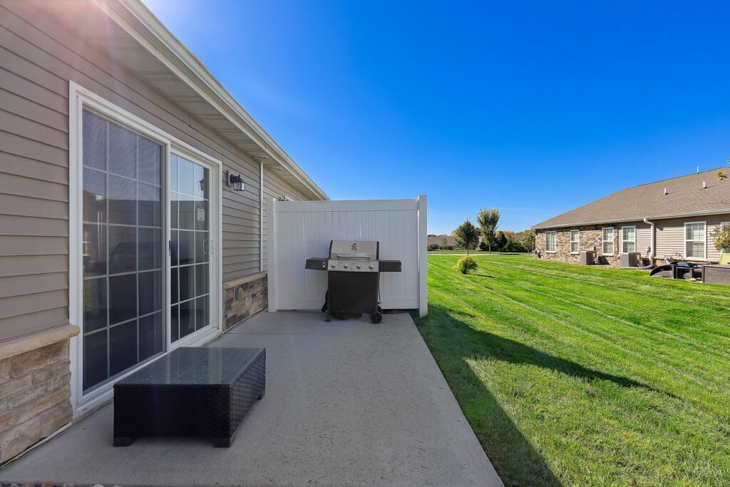 A concrete patio with a wicker table, grill, and a white privacy fence beside a house, overlooking a green lawn under a clear blue sky.