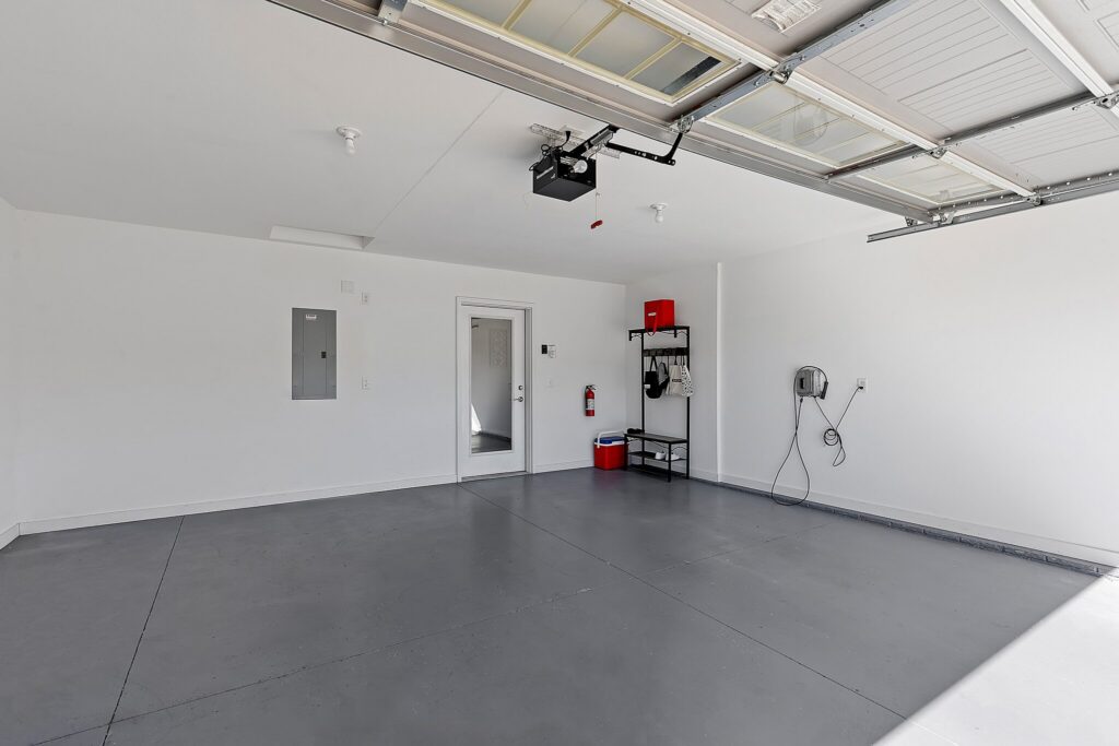 Clean, empty garage with a gray floor, white walls, open garage door, storage shelf, electrical panel, fire extinguisher, and a wall-mounted device on the right.
