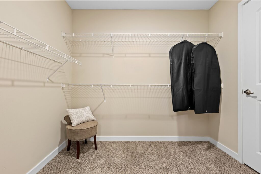 Empty walk-in closet with beige walls, carpeted floor, white wire shelves, a small cushioned stool with a pillow, and two black garment bags hanging on the right.