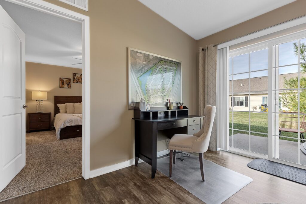 A home office with a dark desk and beige chair sits by a large window; a bedroom with a wooden bed is visible through an open door.
