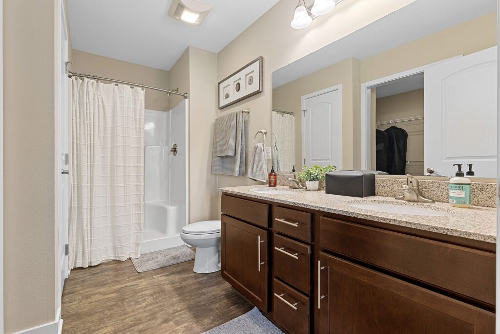 Modern bathroom with a single sink vanity, granite countertop, wood cabinets, toilet, walk-in shower with curtain, towels, and neutral-colored walls and flooring.