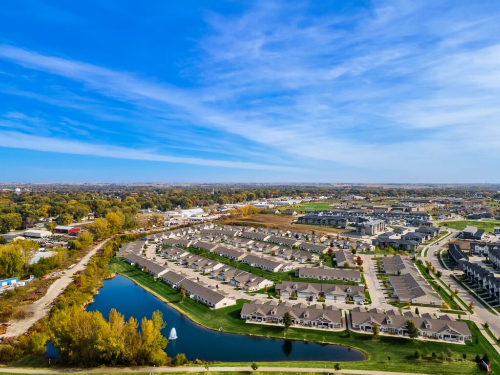 Aerial view of a suburban neighborhood with rows of houses, a small pond, green spaces, and a backdrop of trees under a blue sky.