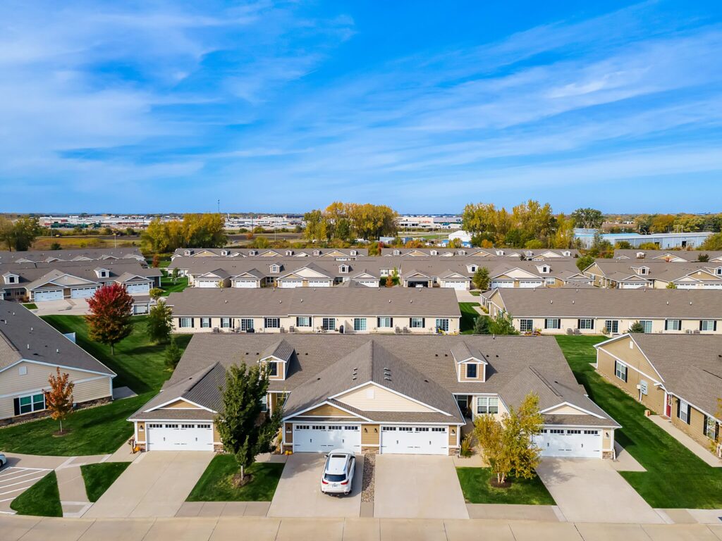 Aerial view of a suburban neighborhood with rows of similar single-story houses, green lawns, and a few trees under a blue sky.