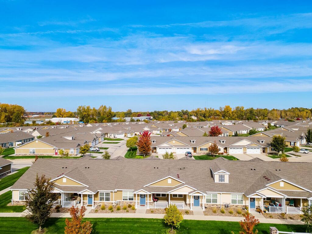 Aerial view of a suburban neighborhood with rows of single-story houses, manicured lawns, trees, and a bright blue sky.