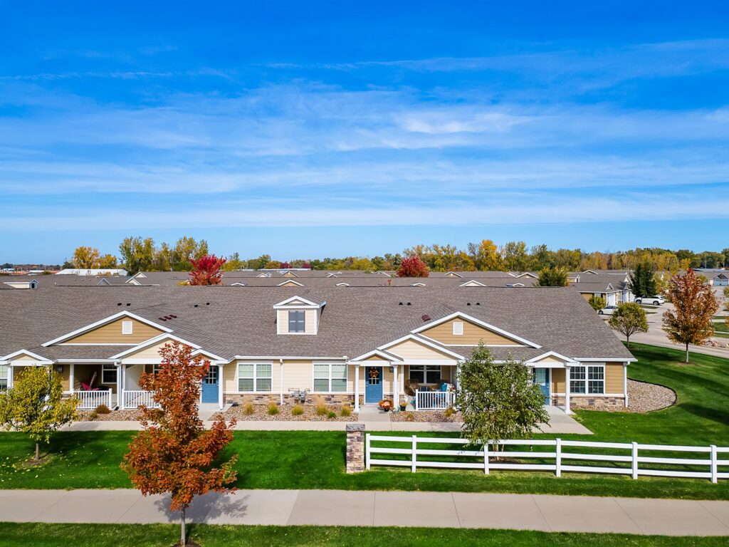 Single-story residential buildings with gray roofs and beige siding, surrounded by green lawns, trees with autumn colors, and a white fence under a clear blue sky.