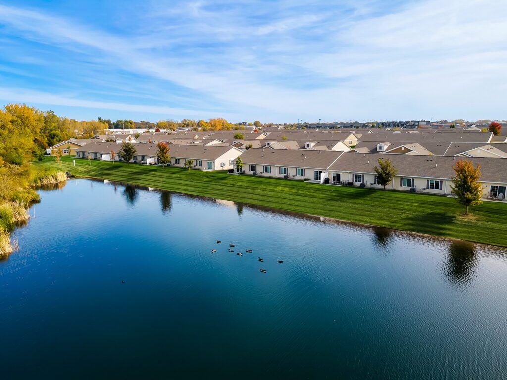 A row of townhouses lines a pond with ducks swimming, surrounded by green grass and autumn trees under a blue sky.
