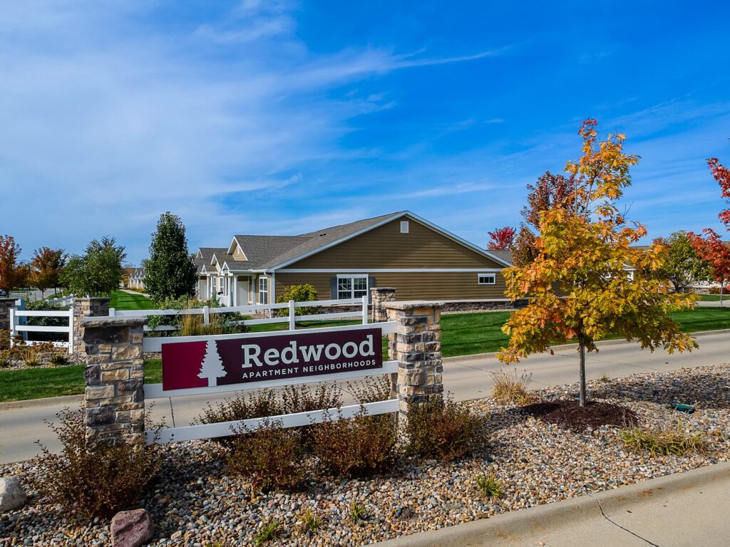 Entrance sign for Redwood Apartment Neighborhoods in front of a single-story residential building, with landscaped stones and autumn trees.