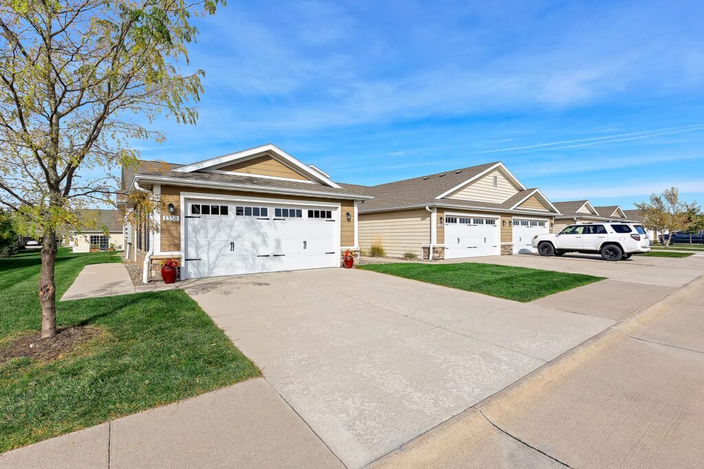 Row of single-story suburban houses with double garages, driveways, green lawns, and a white SUV parked on the right under a clear blue sky.