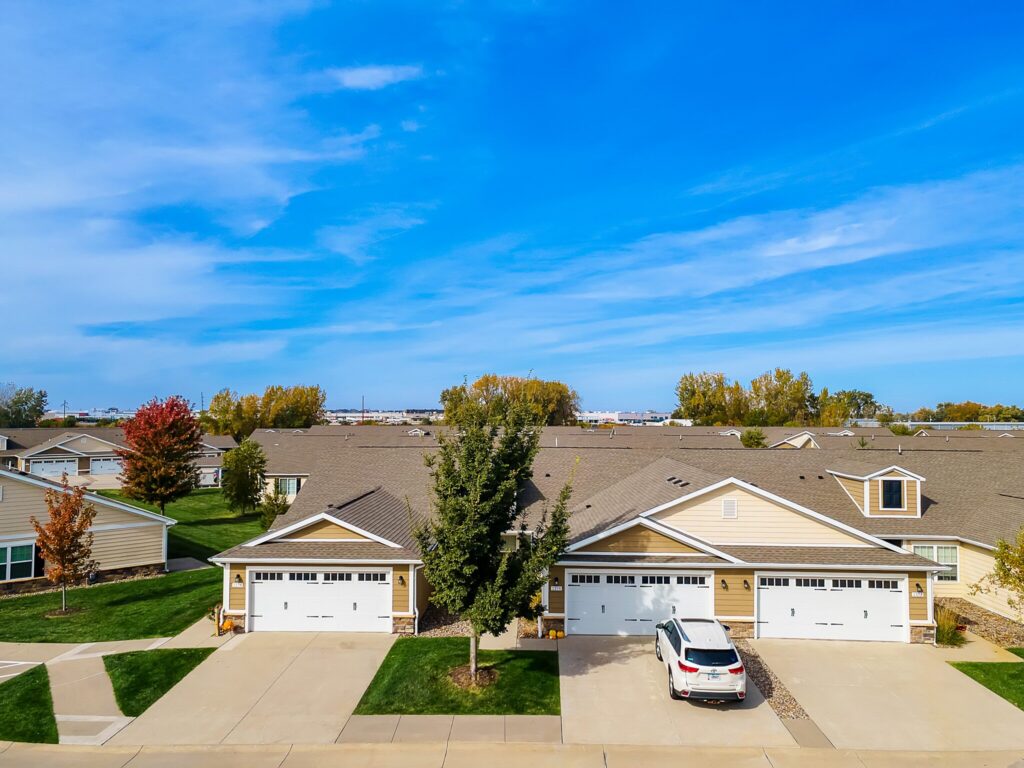 A row of suburban single-story houses with well-kept lawns, driveways, and one white SUV parked in front. Trees with green and autumn-colored leaves are visible. Blue sky overhead.