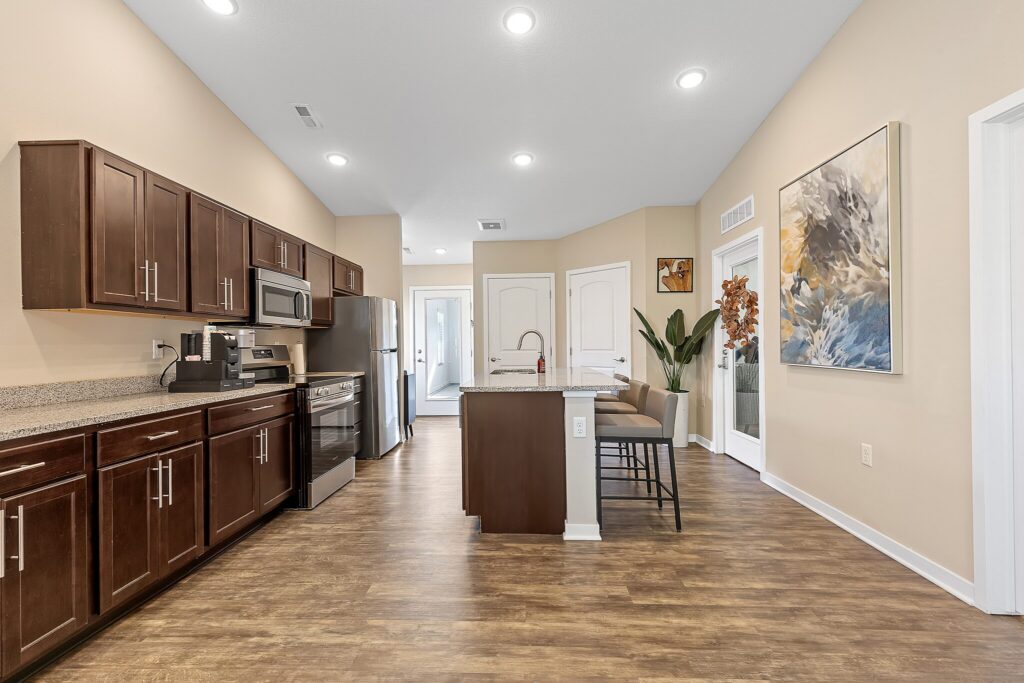 Modern kitchen with dark wood cabinets, stainless steel appliances, a small island with seating, wood floors, neutral walls, and a large abstract painting on the right wall.