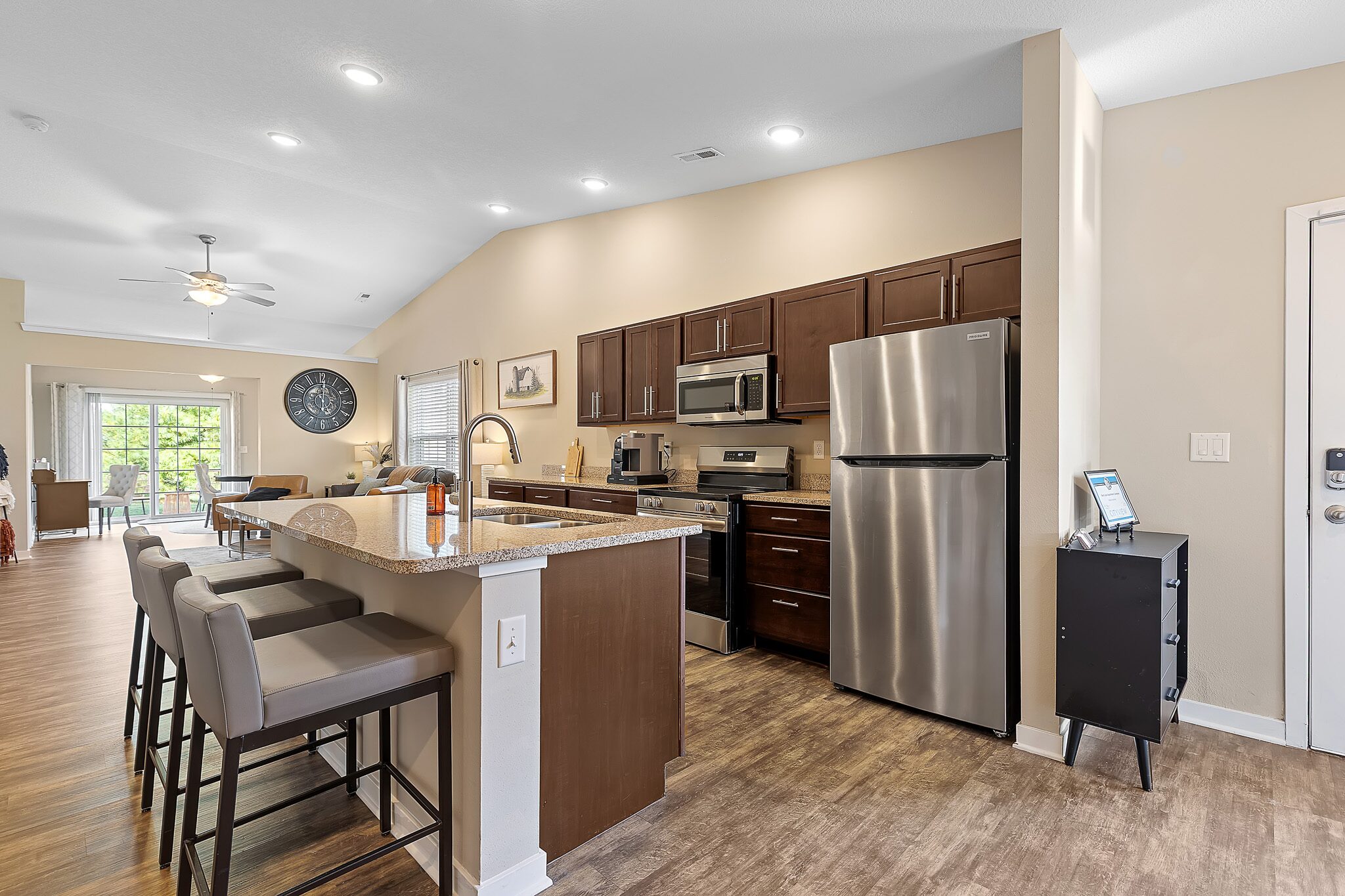 Modern kitchen with stainless steel appliances, brown cabinets, granite countertops, and three barstools at a breakfast bar. Open layout extends to dining and living areas.