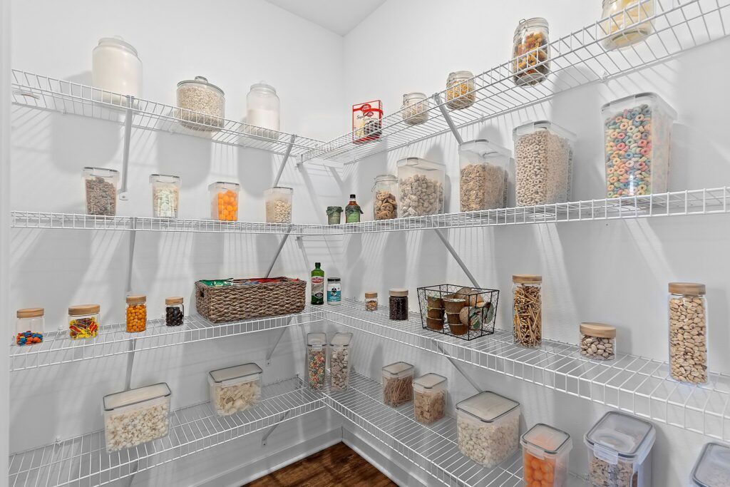 Organized pantry with wire shelves holding clear containers of cereals, pasta, snacks, and dry goods, plus jars, baskets, and small bottles in a white, well-lit room.