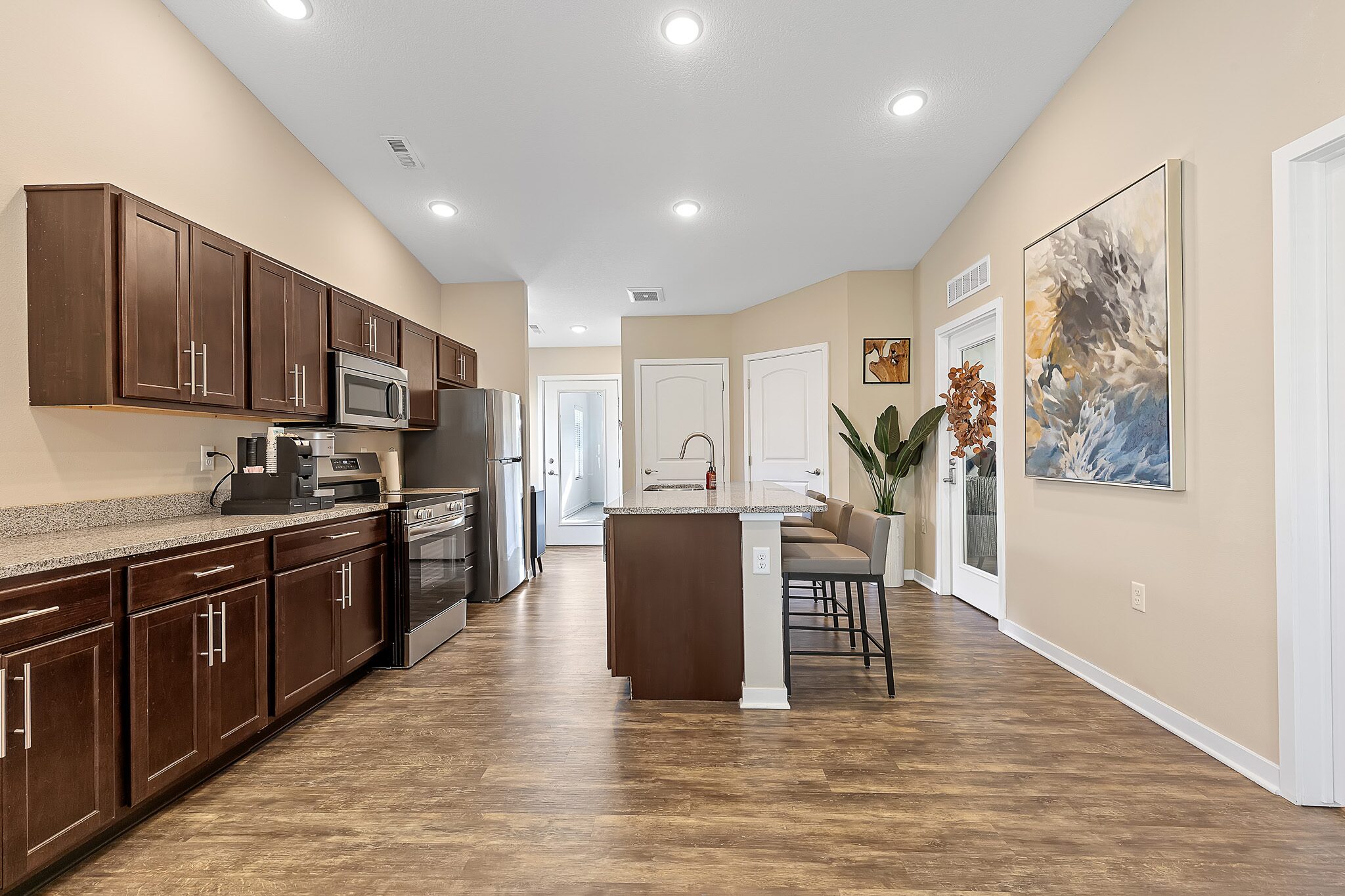 Modern kitchen with dark wood cabinets, stainless steel appliances, a small island with seating, wood floors, neutral walls, and a large abstract painting on the right wall.
