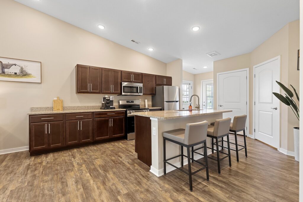 Modern kitchen with dark wood cabinets, stainless steel appliances, an island with three beige barstools, and wood flooring under recessed lighting.