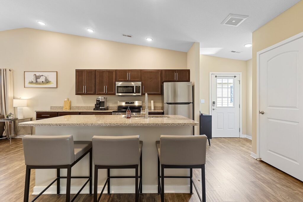 Modern kitchen with granite countertops, three barstools, stainless steel appliances, brown cabinets, wood flooring, and a door leading outside.