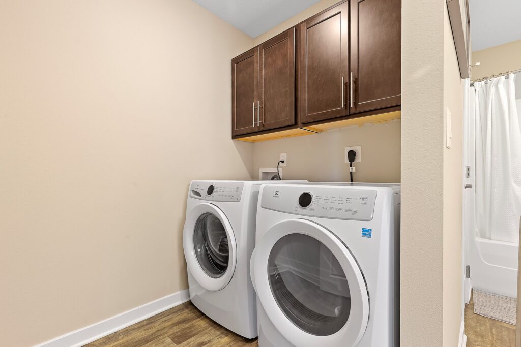 Laundry room with a front-loading washer and dryer, dark wood cabinets above, beige walls, and wood-style flooring. A bathroom with a shower curtain is partially visible on the right.