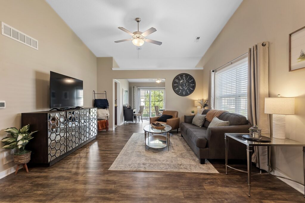 Modern living room with a wall-mounted TV, dark sofa, glass coffee table, large clock, and wood flooring. Dining area visible in the background. Natural light from large windows.