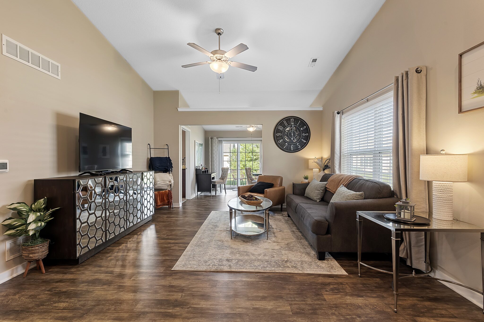 Modern living room with a wall-mounted TV, dark sofa, glass coffee table, large clock, and wood flooring. Dining area visible in the background. Natural light from large windows.