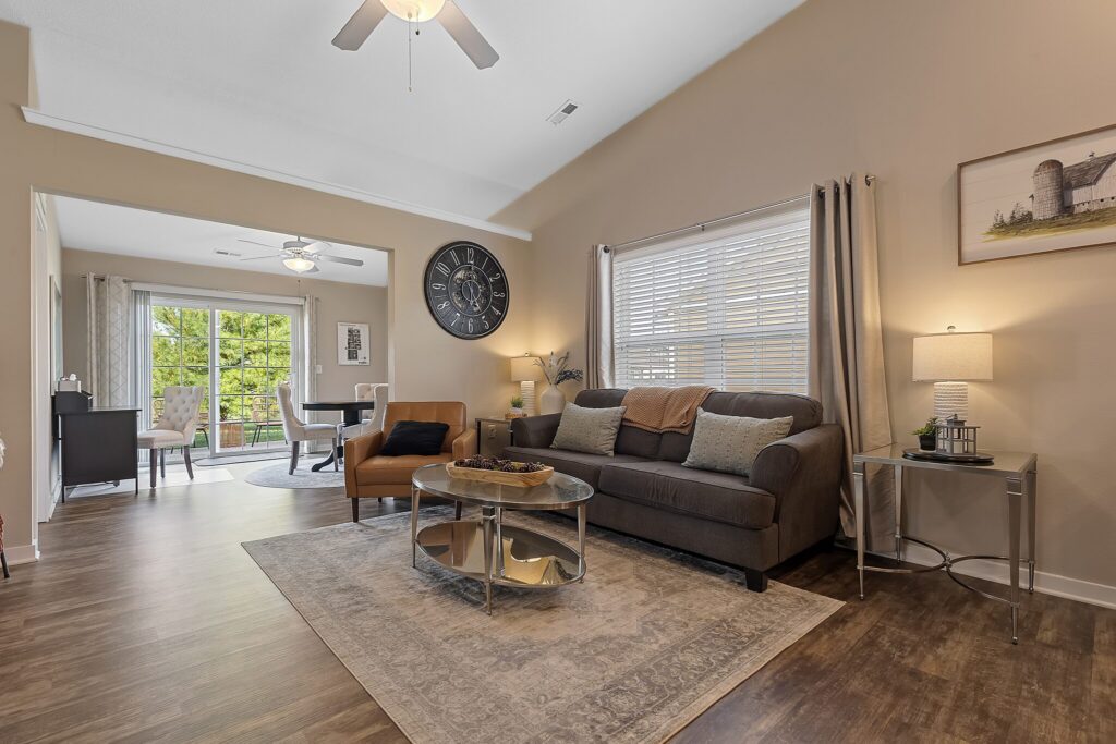 Modern living room with a sofa, armchair, coffee table, area rug, wall clock, lamp, and large window, leading to a dining area with glass doors and wood flooring throughout.