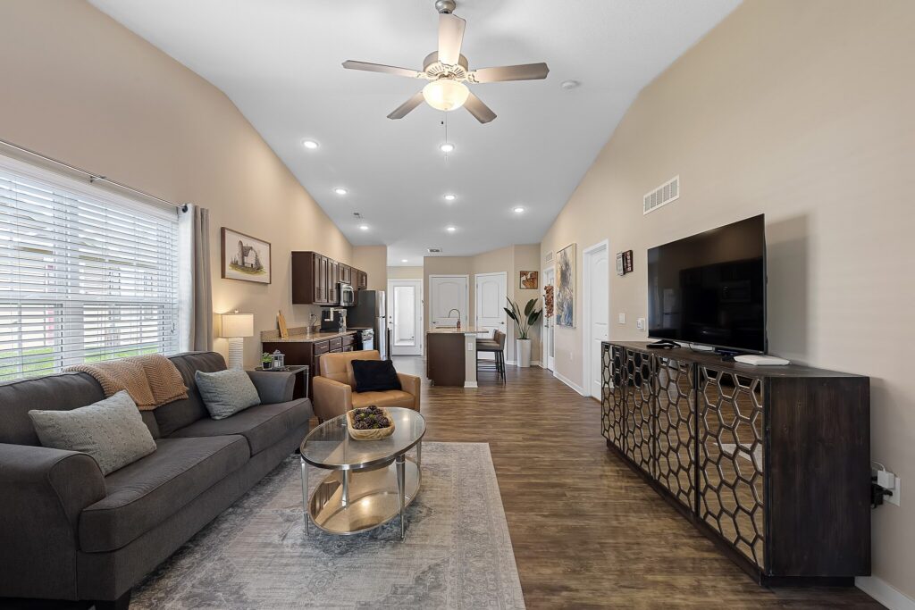 Open-concept living room and kitchen with a ceiling fan, gray sofa, glass coffee table, large TV on a console, and wood flooring. Natural light enters through the window on the left.