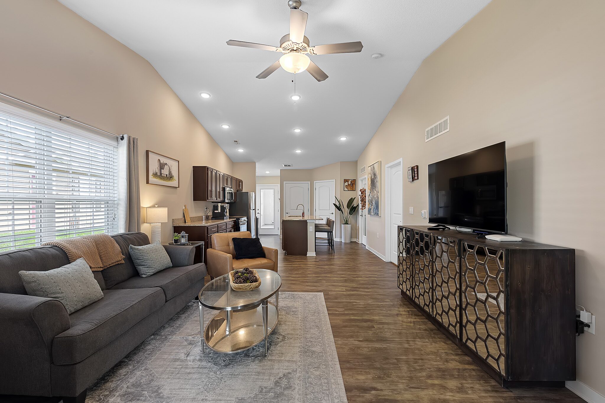 Open-concept living room and kitchen with a ceiling fan, gray sofa, glass coffee table, large TV on a console, and wood flooring. Natural light enters through the window on the left.