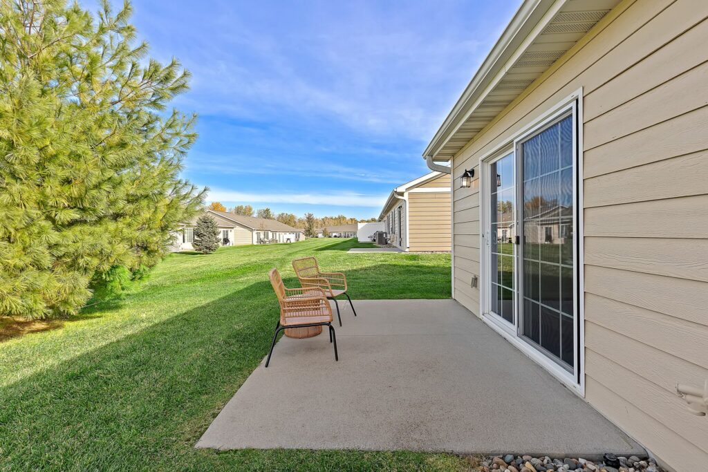 Concrete patio with two wicker chairs beside a beige house, sliding glass door, green lawn, and trees under a blue sky.