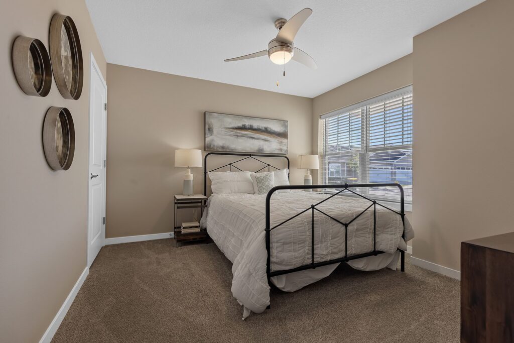 Bedroom with beige walls, a metal bed frame, white bedding, two nightstands with lamps, three round wall decorations, a ceiling fan, and a window with blinds letting in natural light.