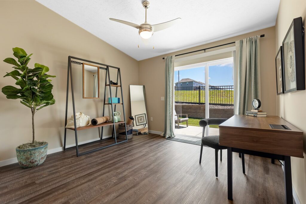 Home office with a desk, chair, potted plant, and leaning mirrors. Sliding glass door opens to a patio with a view of grass and a fence. Ceiling fan and wood floor are visible.
