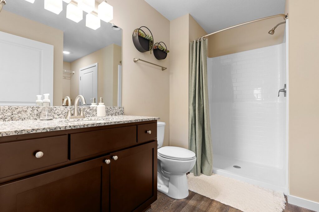 Modern bathroom with a granite countertop, dark wood vanity, toilet, walk-in shower with a green curtain, wall-mounted planters, and beige walls.