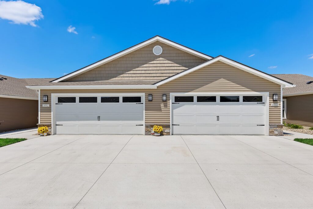 A beige house with two double garage doors, four outdoor lights, concrete driveway, and potted yellow flowers by each garage door, under a clear blue sky.