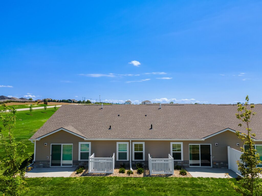 Single-story duplex with gray siding, white trim, two small porches, and a well-maintained green lawn under a clear blue sky.