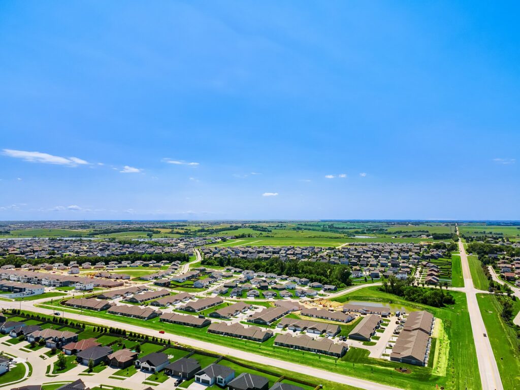 Aerial view of a suburban neighborhood with rows of houses, green lawns, trees, and roads under a clear blue sky.