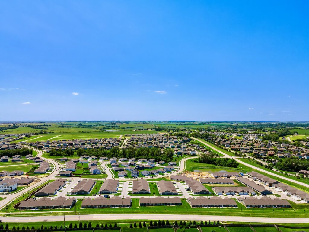 Aerial view of suburban housing development with rows of houses, green lawns, roads, and open fields under a clear blue sky.