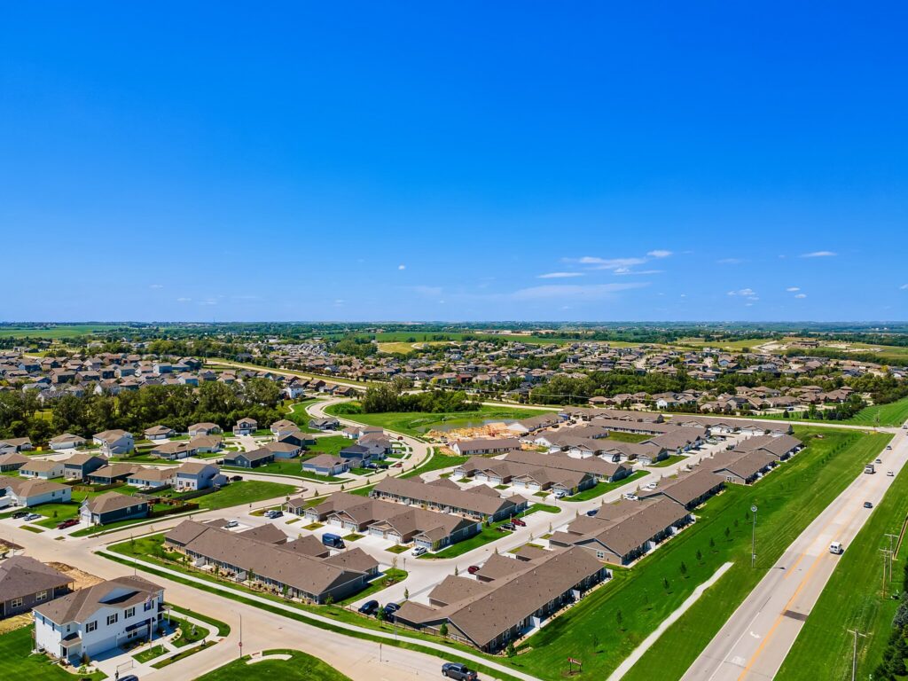 Aerial view of a suburban neighborhood with rows of houses, roads, and green lawns under a clear blue sky.