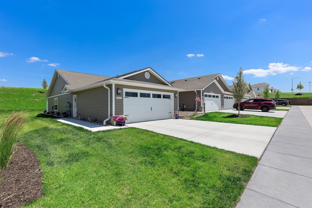 Single-story suburban homes with attached garages, driveways, and well-kept lawns on a sunny day. A red SUV is parked in one driveway; an American flag is displayed on one house.